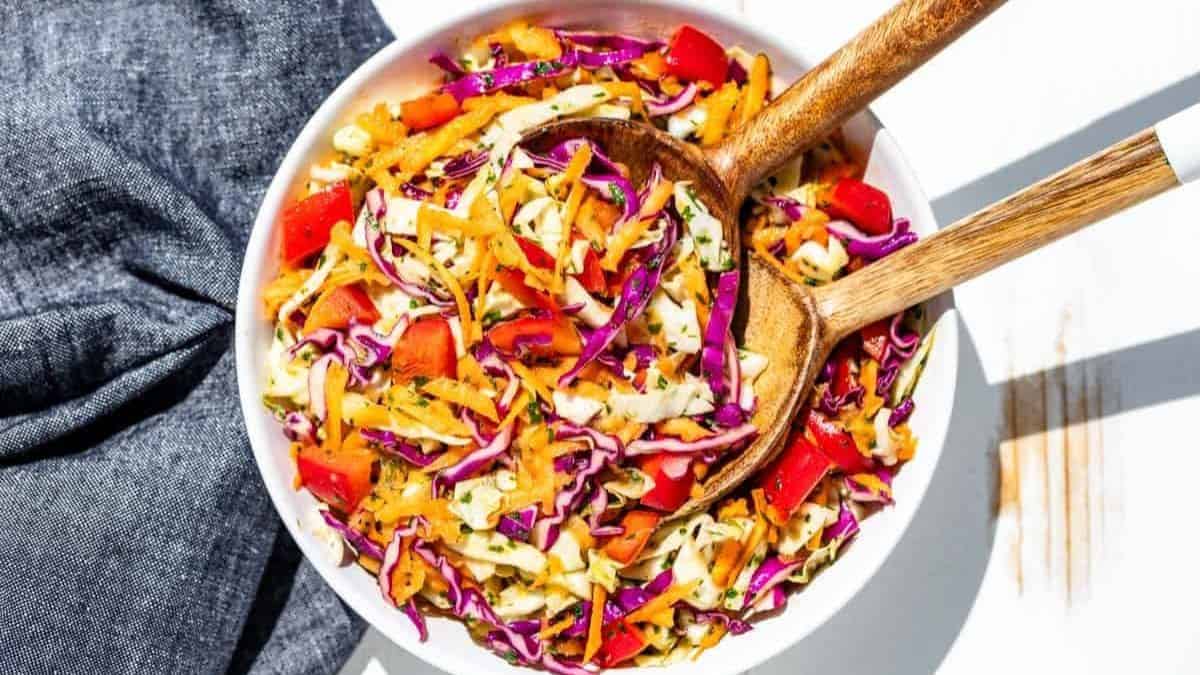 A top shot view of a white ceramic bowl with two wooden serving spoons, filled with Cilantro Lime Slaw. 