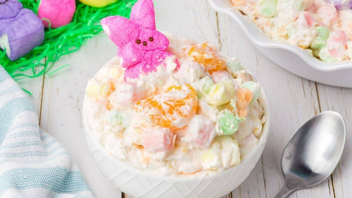 A serving of Easter Fruit Fluff sitting on a white ceramic bowl, a spoon can be seen on its sides. 