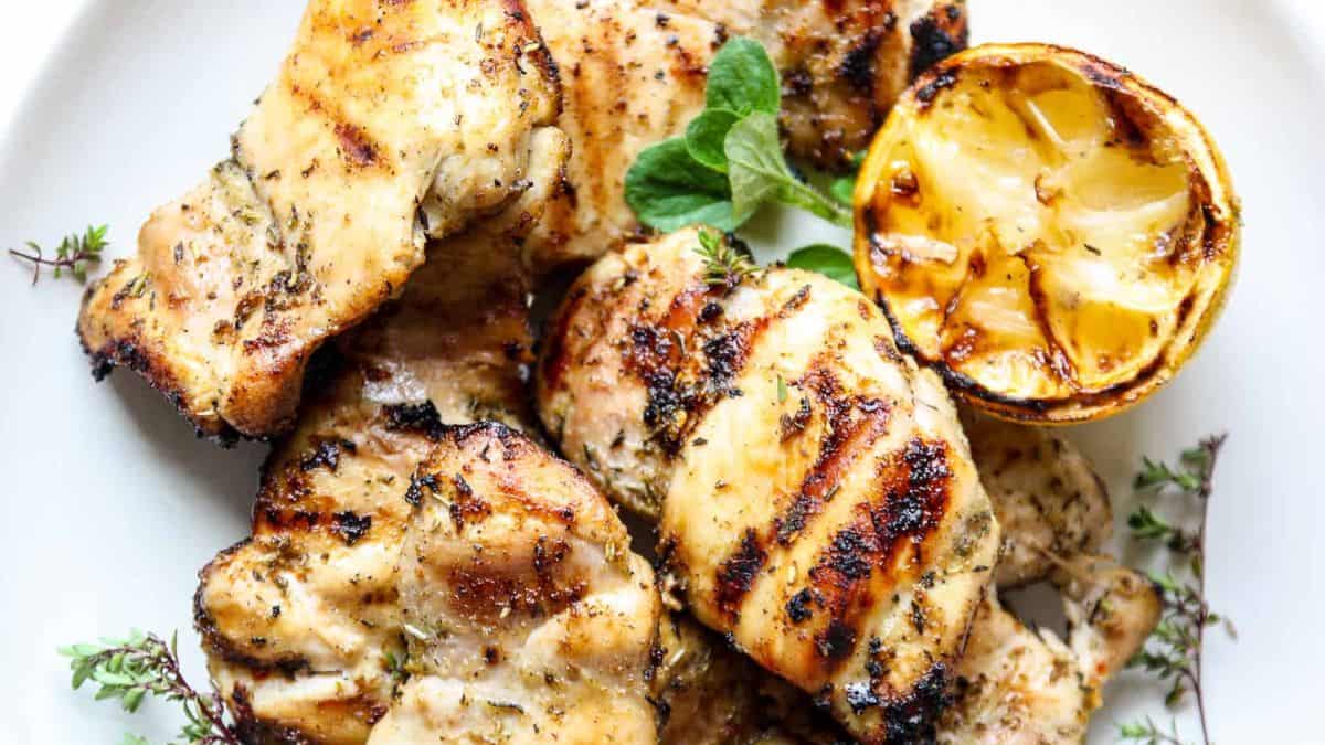 A close-up shot of Grilled chicken set on a plate with a half of grilled lemon and some herbs.