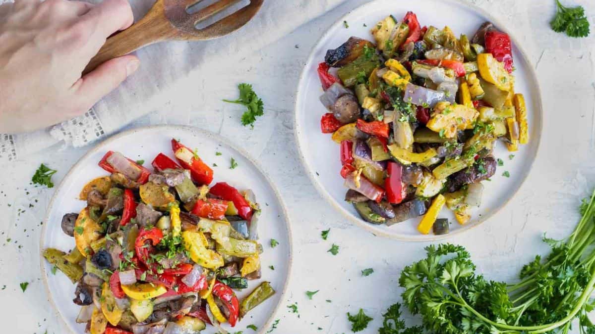 A hand holding a wooden ladle, and next to it are two plates filled with Italian Oven-Roasted Vegetables. 