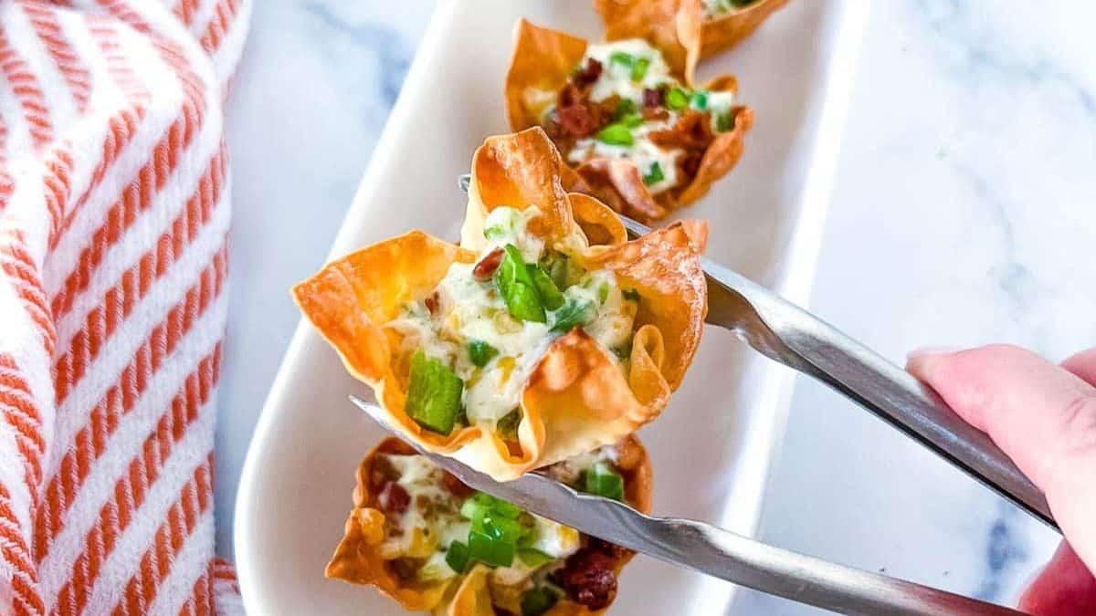 A close-up shot of a Jalapeño Popper Wontons held using a thong, and the other couple of pieces can be seen from the background set on a plate. 