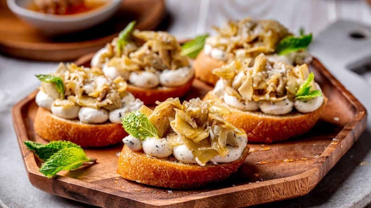 A wooden plate with four pieces of  Crostini topped with Marinated Artichokes.