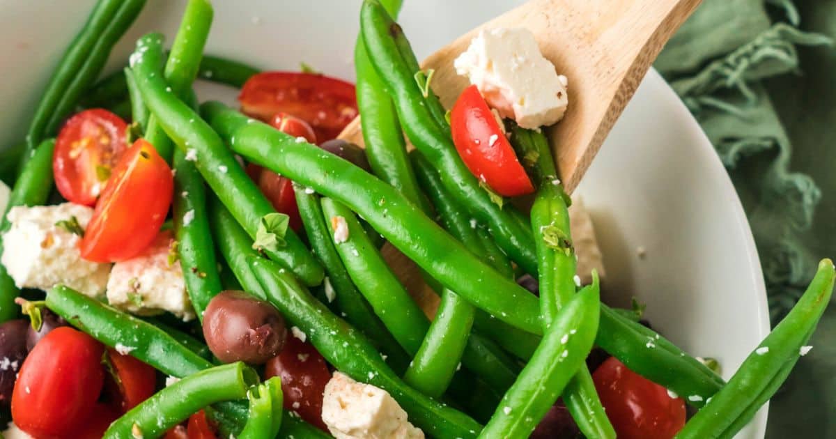 A bowl of green beans with tomatoes and some blocks of feta cheese