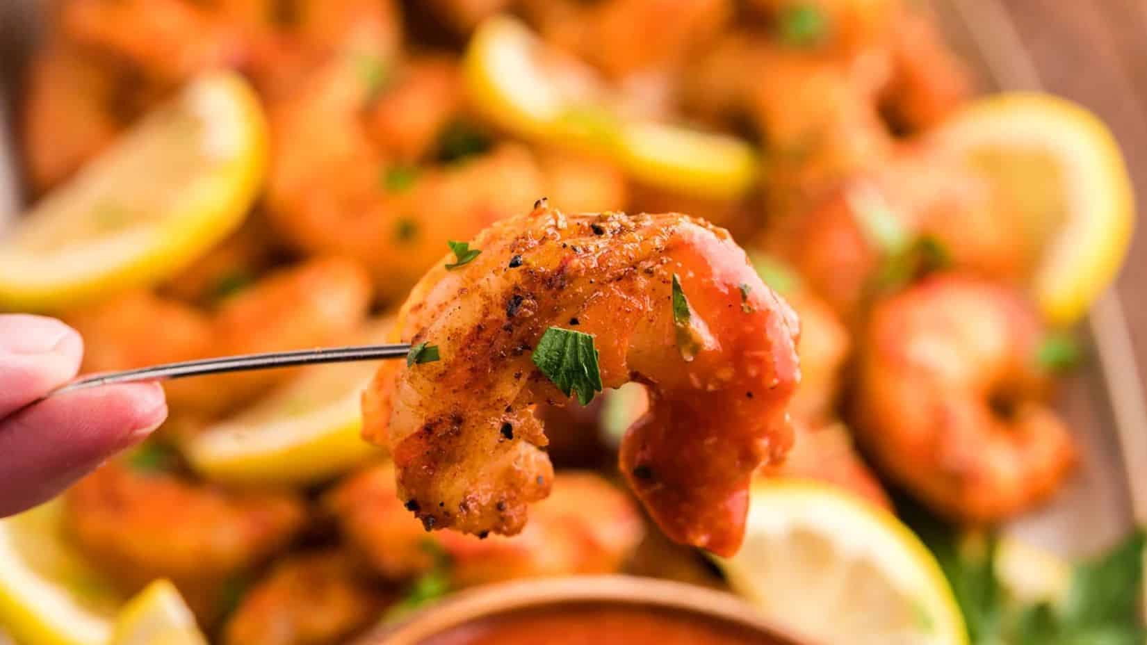 A close-up shot of a single smoked shrimp poked with a stick held by two fingers, a blurry background of the rest of the shrimp and lemon slices. 