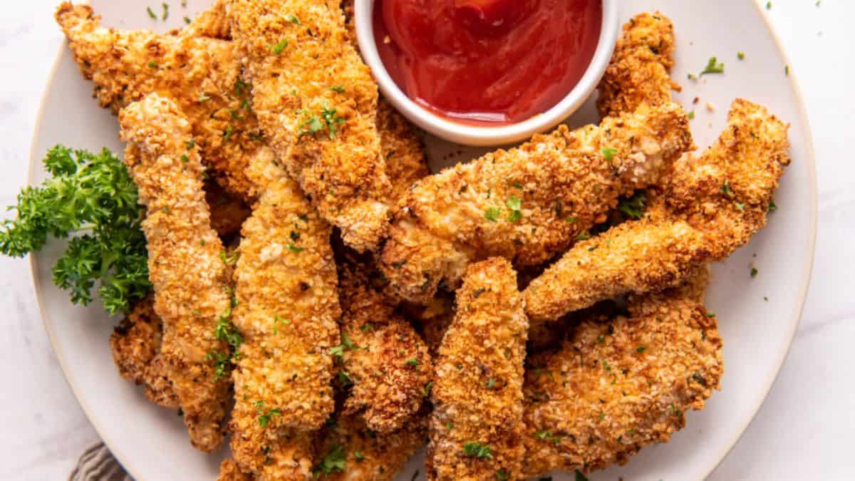 Top shot of a plate with a couple of chicken strips with a dip on the side. 