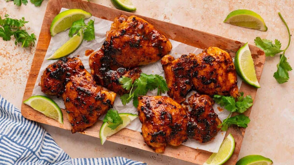 Top shot of a wooden board with four pieces of baked boneless chicken thighs on top of it, with some slices of lime on the sides.
