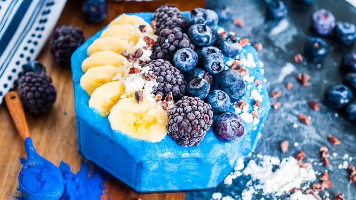 A clear bowl filled with blue colored smoothie and topped with slices of banana, blueberries, and blackberries.