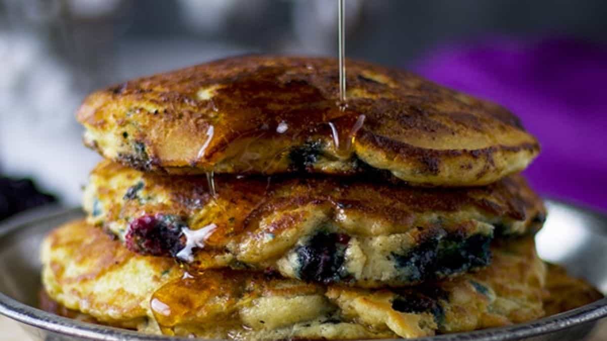 Three stack of pancakes with blueberries set on a plate while being drizzled with maple syrup. 