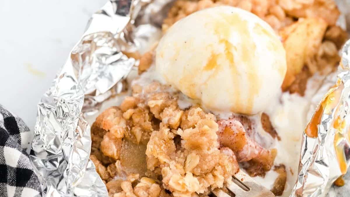 A close-up shot of Campfire Apple Crisp Foil Packets topped with a scoop of vanilla ice cream and some drizzle of caramel.