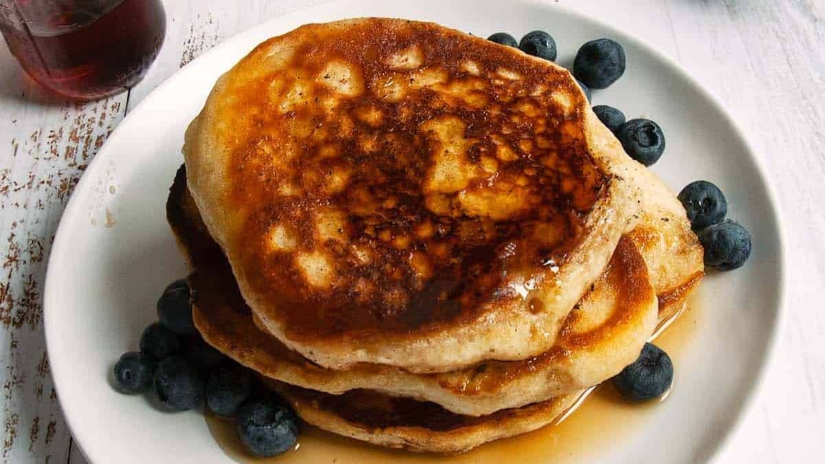 Three-stack of pancake set on a plate, with blueberries and maple syrup.
