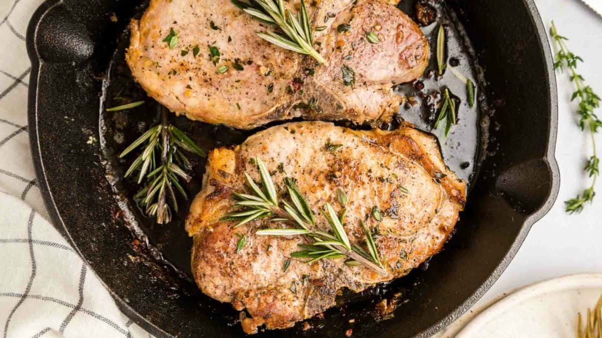 Two pieces of porkchop cut cooked on a cast iron with a few pieces of rosemary leaves.