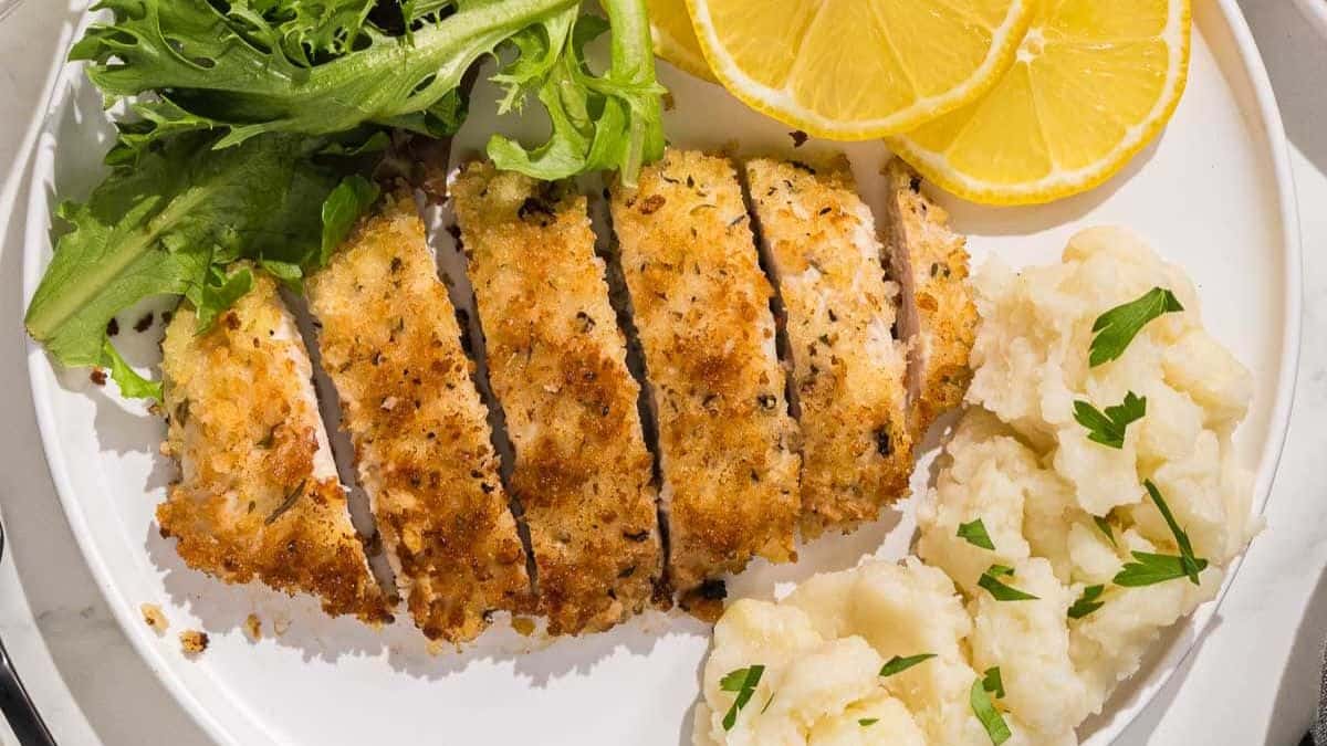 A plate with a piece of Italian Cutlet, mashed potato and salad on the side and some slices of lemon. 