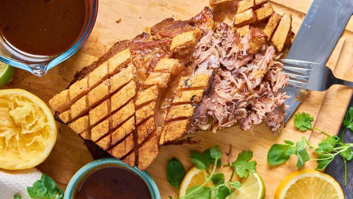 Top shot of a cooked Cuban pork shoulder set on a wooden chopping board with a knife, fork, some sauce on a container and some lemon slices. 