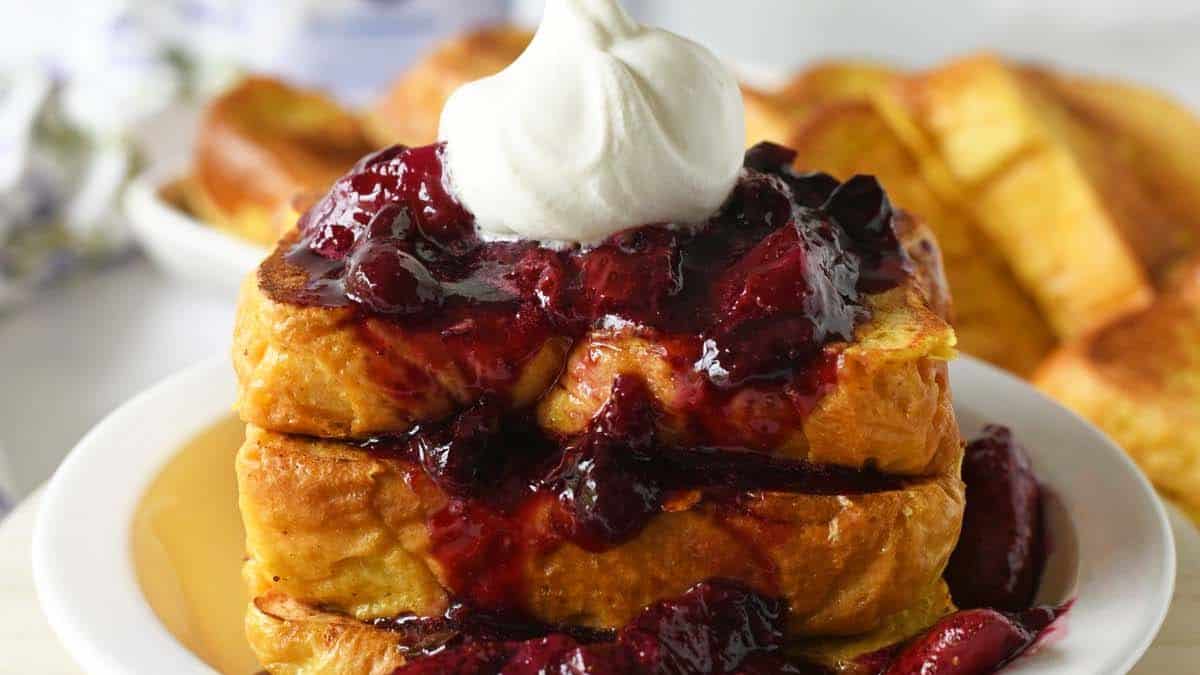Three stack French toasts set on a plate, with blueberry compote and some cream. 