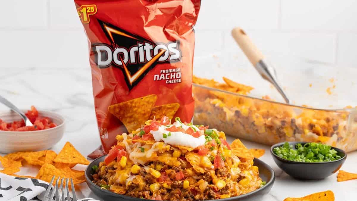 A bag of Doritos chips, and a plate of Doritos casserole, on the side a few pieces of Doritos chips is scattered, a small bowl of salsa, a clear baking dish with the rest of the casserole. 