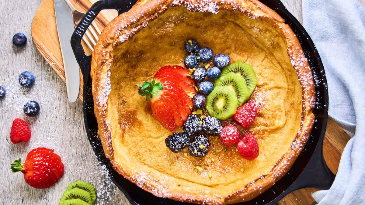 Top shot of a cast iron with German pancakes, topped with slices of fresh fruits, namely: kiwi, strawberry, blueberry, blackberry, raspberriy.