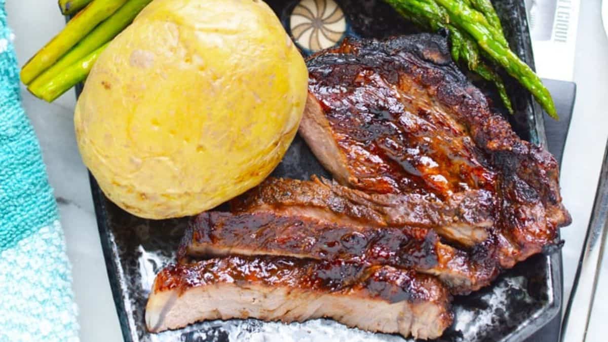 A close-up shot of grilled strip steaks with a whole boiled potatoes and some asparagus on the side. 