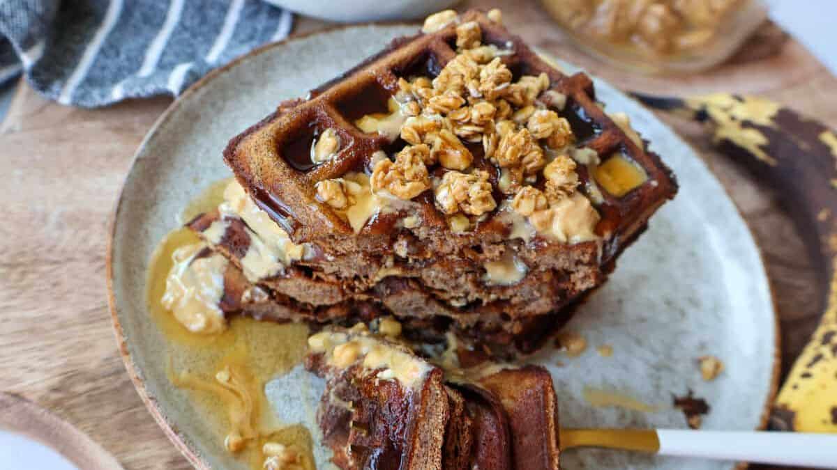 A stack of waffles set on a plate topped with granola and drizzled with syrup while being cut using a fork.