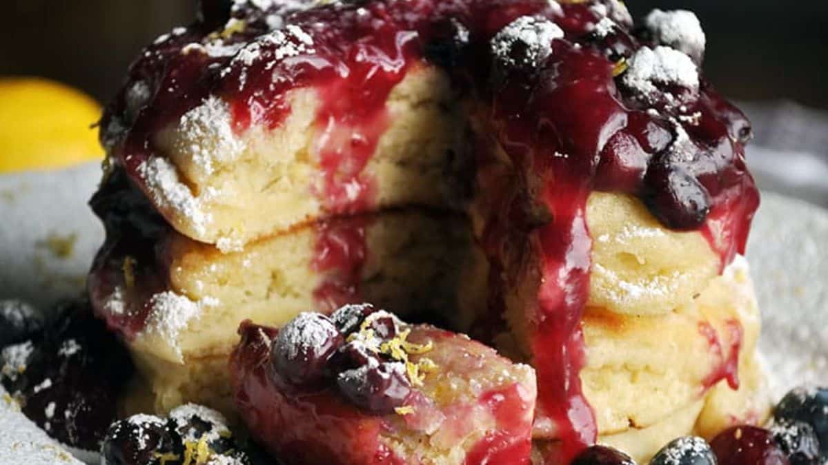 A close-up shot of three tier stack pancake, set on a plate with blueberry sauce and fruits then dusted with powdered sugar. 