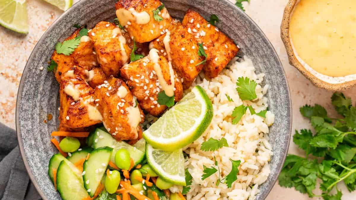 Top shot of a bowl filled with rice and salmon bites and drizzled with bang bang sauce, some slices of lime on top of the rice and cilantro leaves.