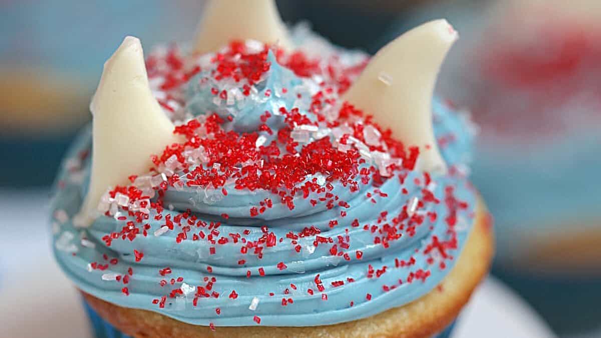 Close-up shot of a cupcake with blue icing and red sprinkles and while shark candies on top.