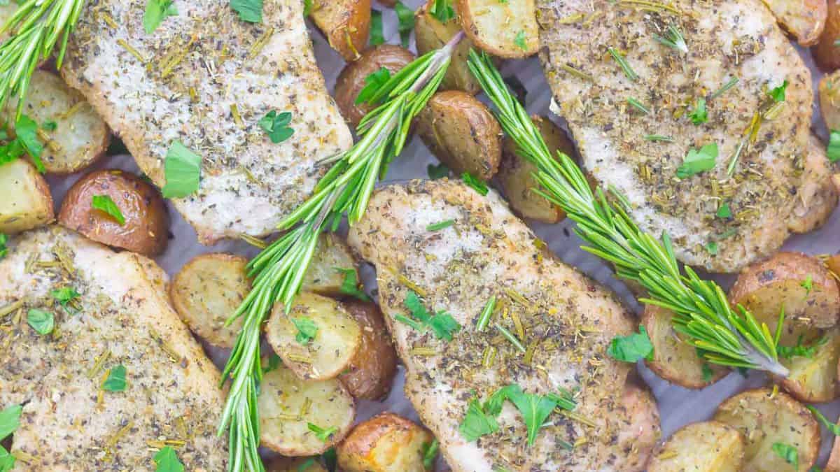 Top shot of a baking tray lined with parchment set with a couple of pieces of halved potatoes and rosemary leaves and four pieces of porkchop cuts. 