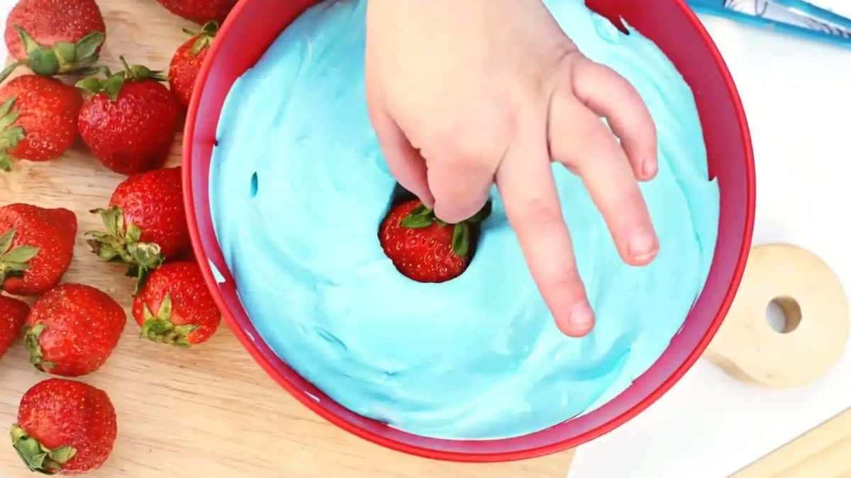 Bowl filled with blue dip and a hand holding a strawberry being dipped to the mixture.
