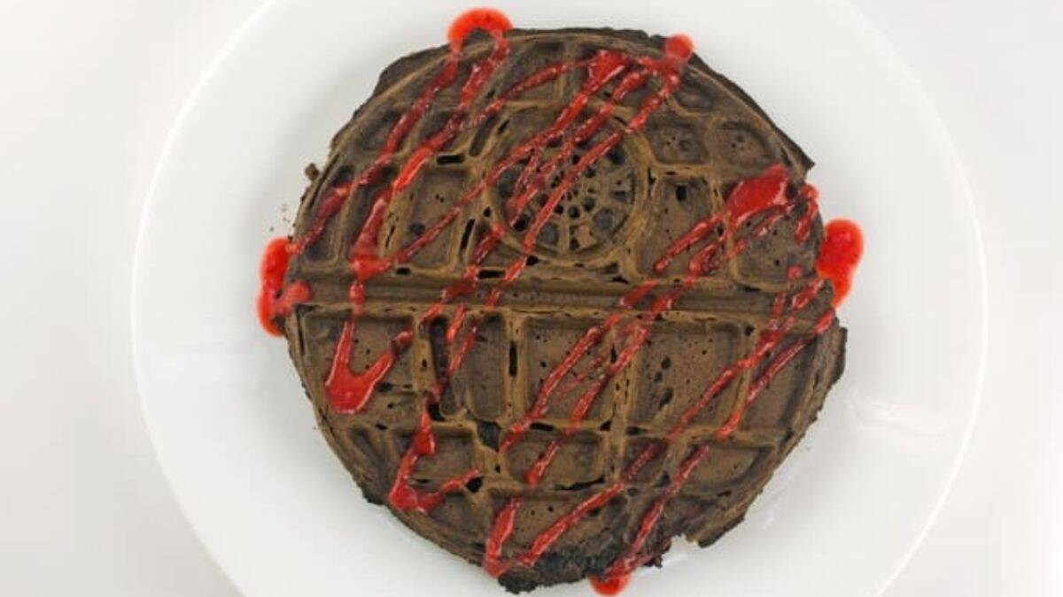 A photo of a chocolate waffle design with the Death Star set on a plate, with a drizzle of Strawberry Sauce.  
