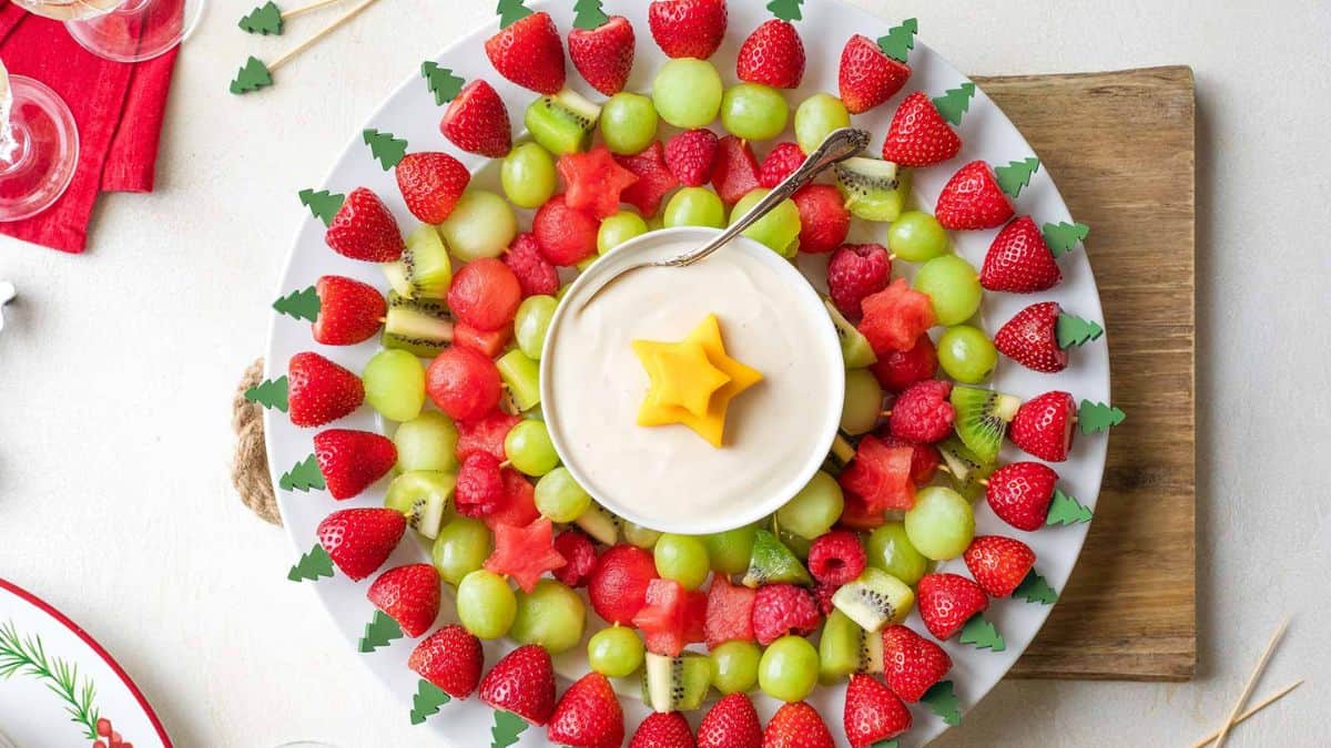 A platter on a wooden board filled with fruit kabobs made with strawberries, kiwis, grapes, and watermelon. In the middle of the platter a bowl of cream. 