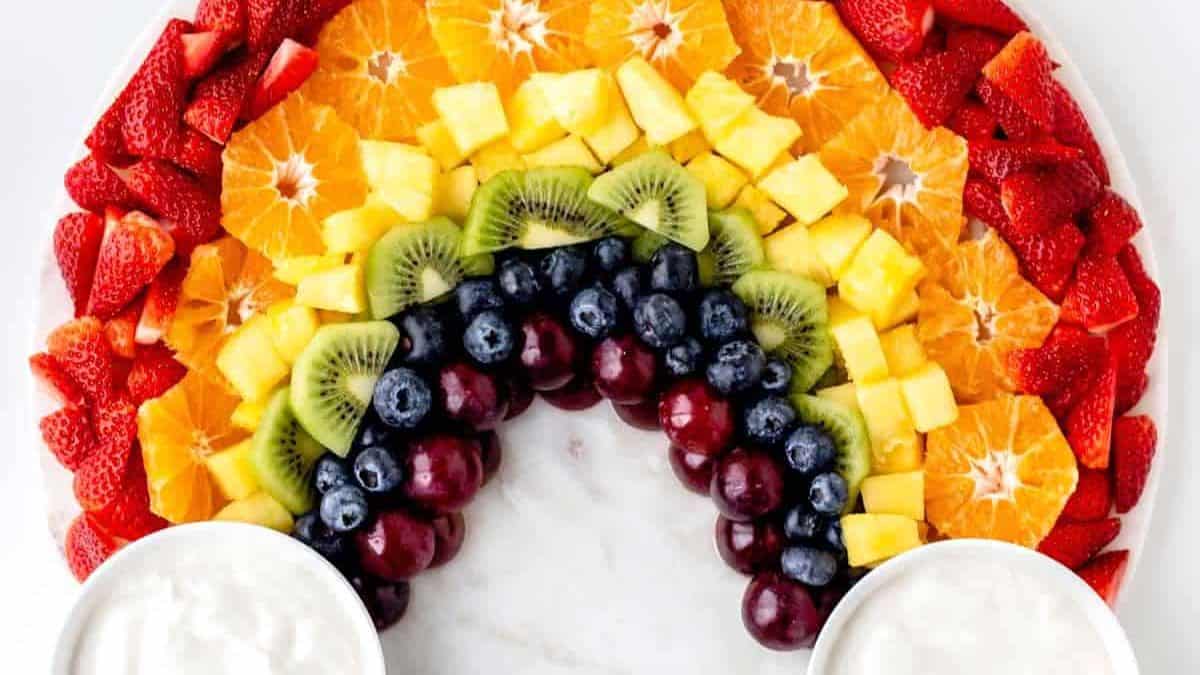 Slices of strawberries, oranges, pineapples, kiwis, blueberries, and grapes all formed like a rainbow and two bowl of cream. 
