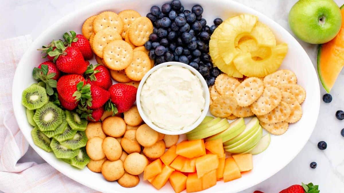 A platter filled with strawberries, slices of kiwi, blueberries, slices of pineapples, slices of green apples, cantaloupe cubes, and different type of crackers, in the middle is a bowl of cream. 