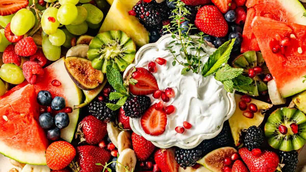 Fruit Charcuterie board with kiwi, blueberries, watermelon, strawberries, figs, banana slices, pineapples, grapes and a bowl of cream. 