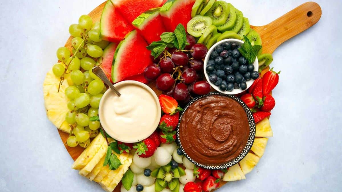 A round board filled with various fruits and dips: slices of kiwi, grapes, blueberries in a small bowl, strawberries halves, slices of pineapples, chocolate dip and cream dip. 