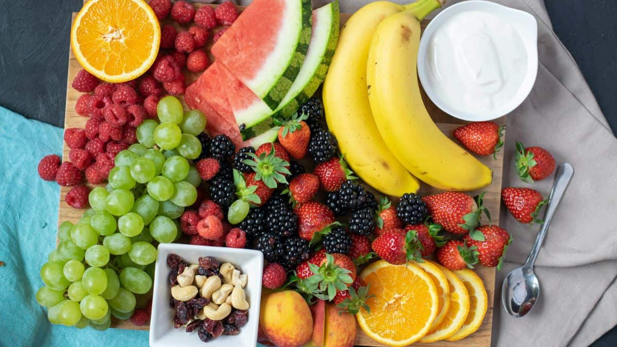 A board filled with various fruits: bananas, oranges, raspberries, blackberries, peaches, grapes, watermelon, a bowl filled with various nuts in a small bowl, a bowl of cream. 