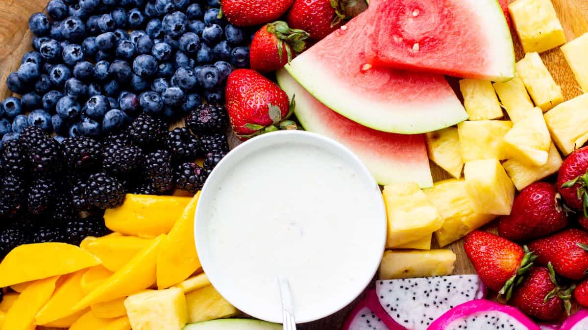 A board filled with different fruits: blueberries, blackberries, slices of mangoes, strawberries, cubes of pineapples, slices dragon fruit, wedges of watermelon and a small bowl of cream in the middle. 