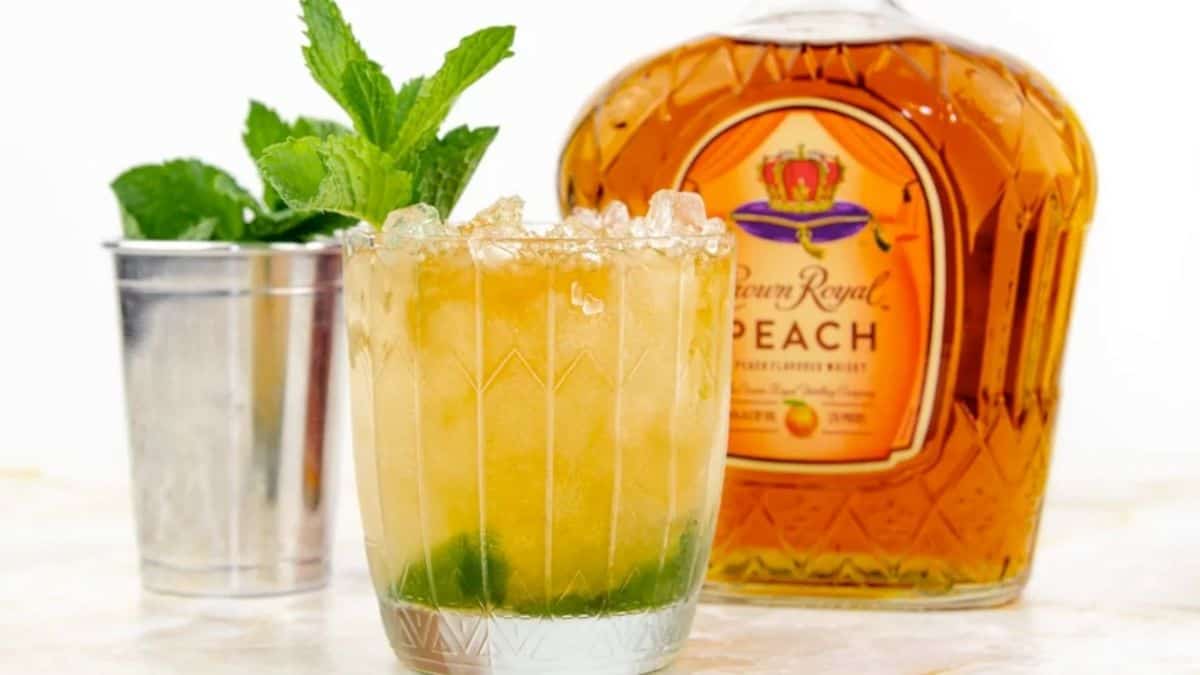 A glass of Peach Mint Julep with a few pieces of mint leaves and a bottle of Crown Royal Peach on the background, and a small bucket of ice. 
