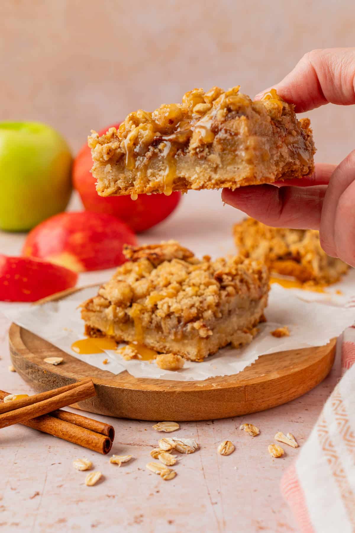 Hand holding a caramel apple pie bar with a bite taken out, showing the crumble topping and gooey apple filling, with another bar on a wood board below