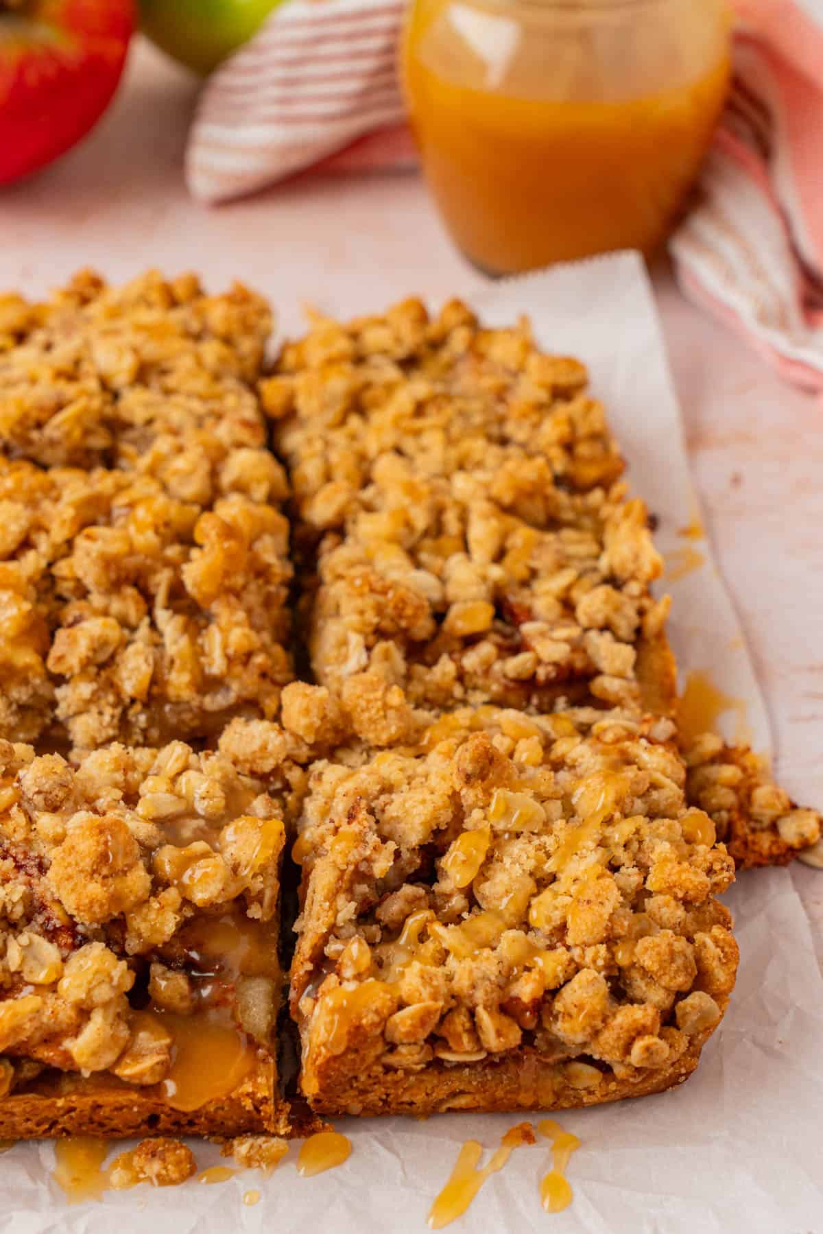 Close-up of sliced apple pie bars in the pan with a jar of homemade salted caramel sauce and fresh apples in the background