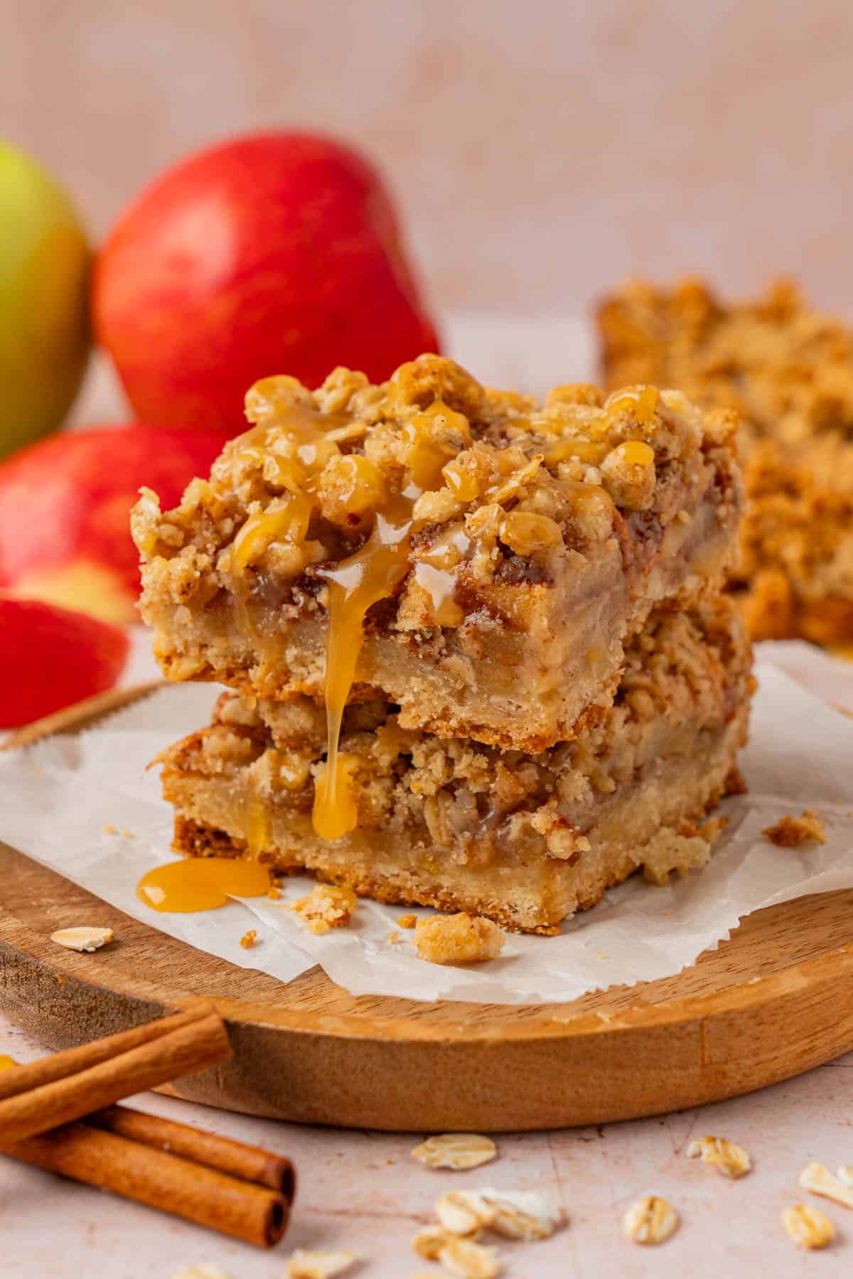 Two stacked salted caramel apple pie bars on parchment paper with caramel sauce dripping down the side, fresh apples and cinnamon sticks in the background