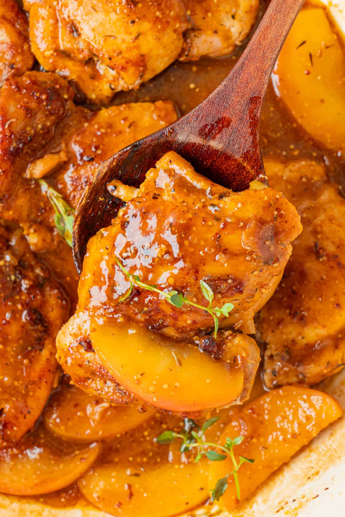 Wooden spoon lifting an apple cider glazed chicken thigh with a tender apple slice and fresh thyme.