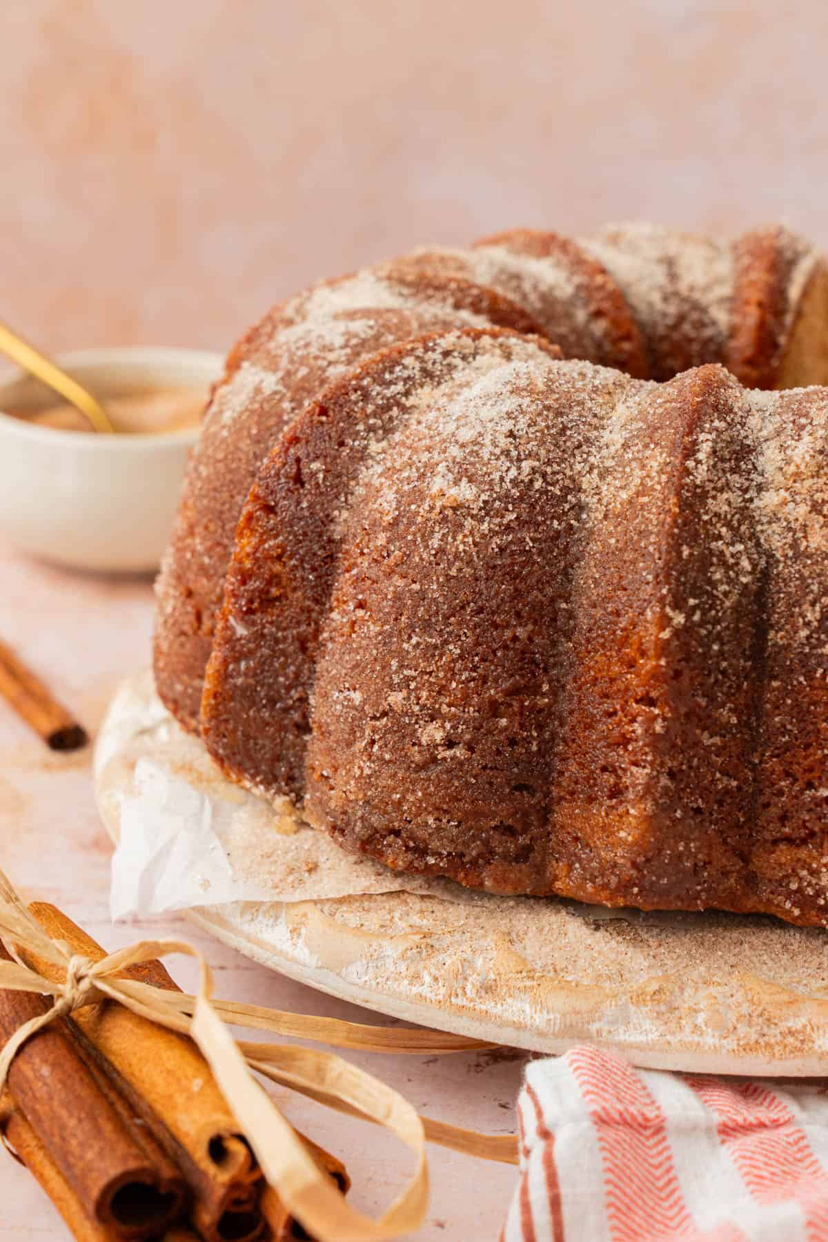 Side view of apple cider donut cake with a sparkly cinnamon sugar crust on a white cake stand next to cinnamon sticks