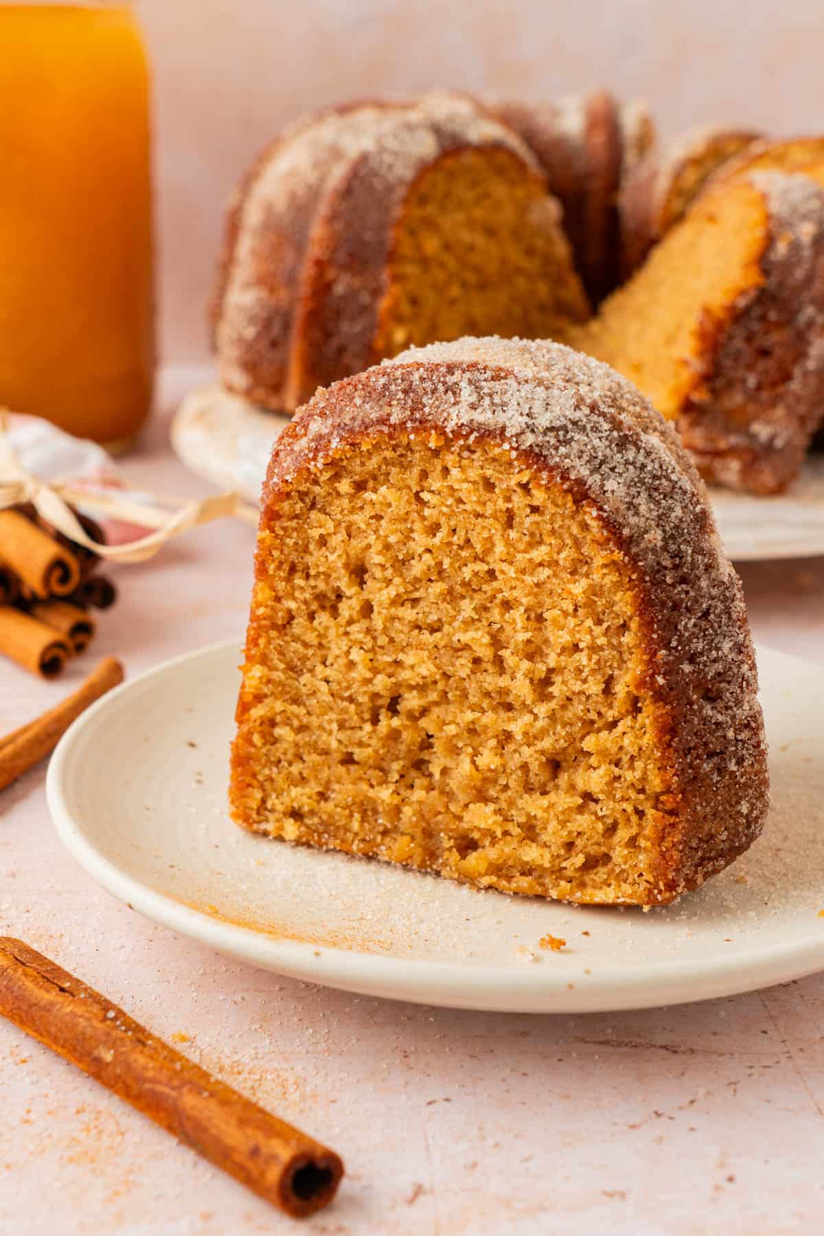 Slice of flavorful apple cider doughnut cake on a plate with a glass of apple cider and the sliced Bundt cake behind it