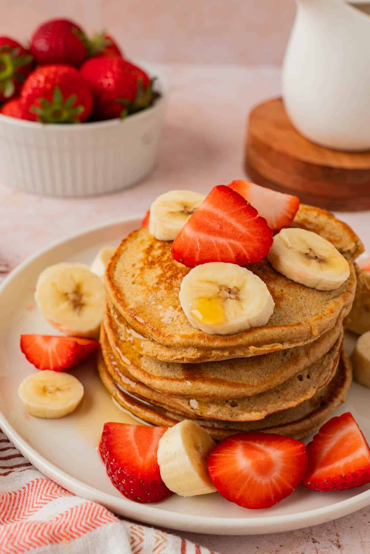 A stack of banana oatmeal pancakes topped with sliced bananas, fresh strawberries, and maple syrup on a white plate.