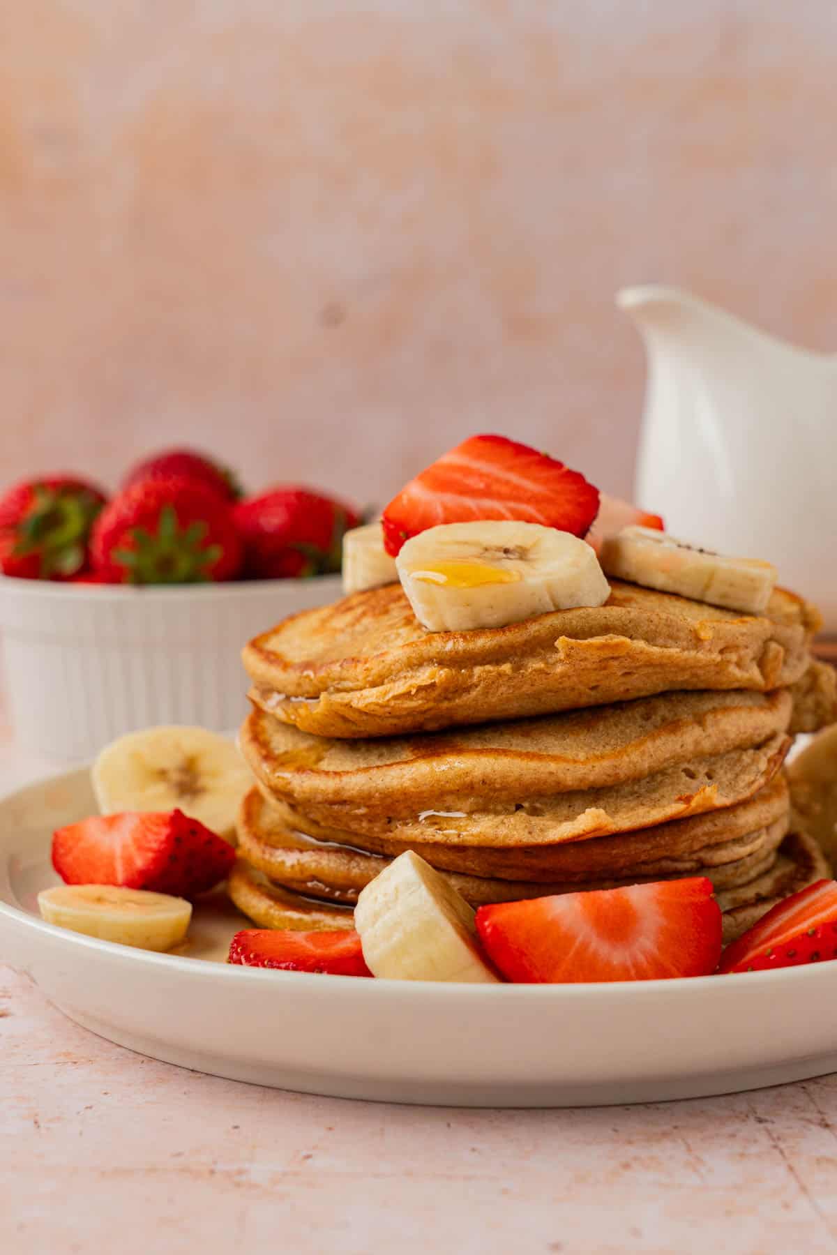 Side view of fluffy banana and oatmeal pancakes stacked high with banana slices and strawberries, a syrup pitcher in the background.
