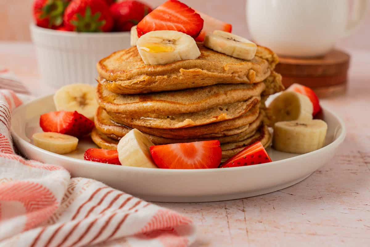 Easy oatmeal banana pancakes stacked on a white plate with fresh strawberries, banana slices, and a striped kitchen towel.