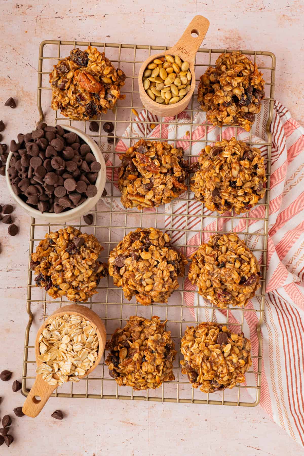 Overhead view of banana oatmeal breakfast cookies on a wire rack surrounded by bowls of chocolate chips, pumpkin seeds, and old fashioned oats