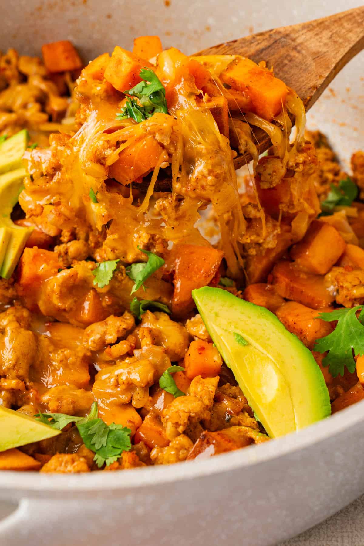 Wooden spoon lifting a cheesy bite of turkey and sweet potato skillet with tender sweet potato cubes and fresh cilantro.