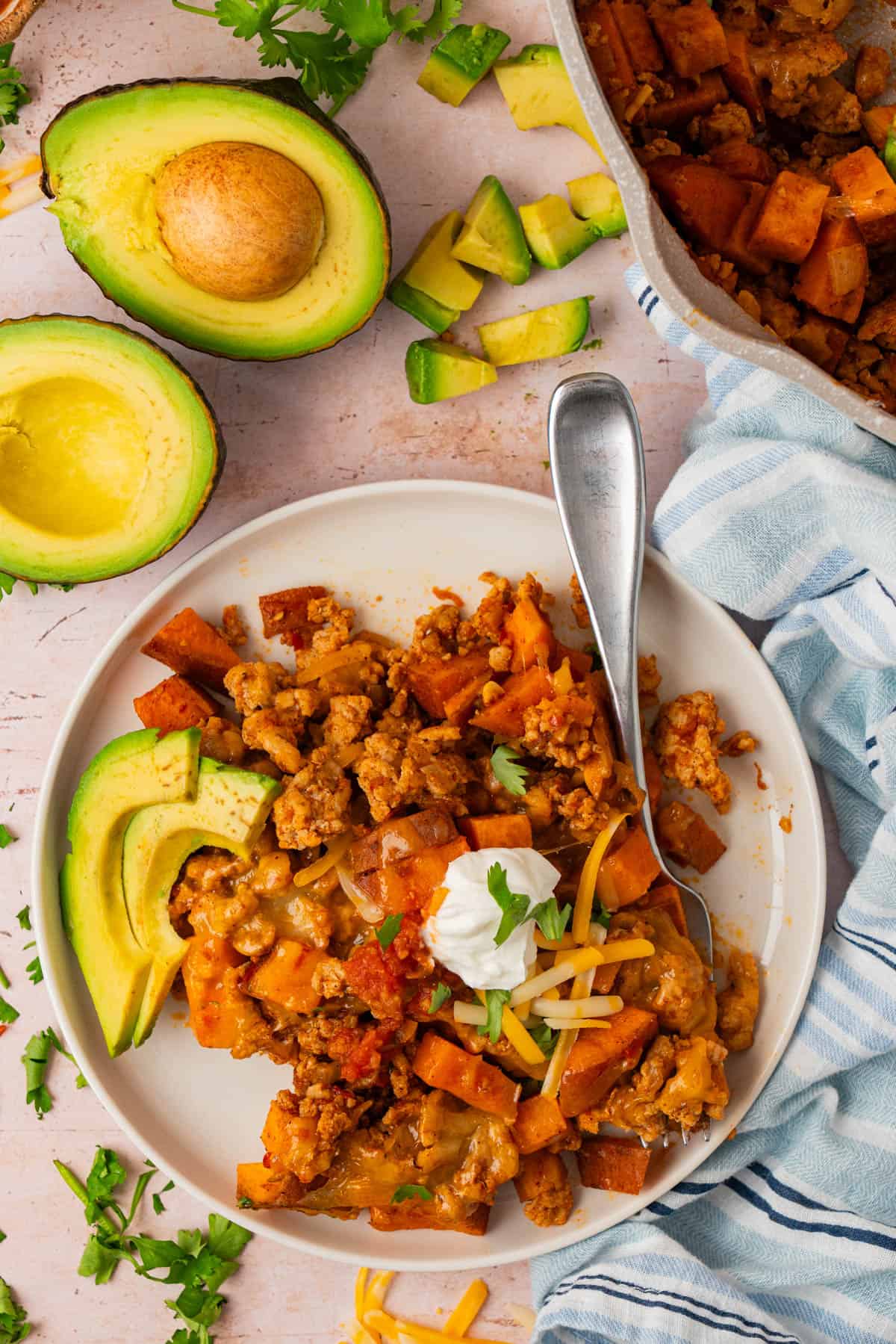 Serving of sweet potato and ground turkey skillet with avocado, Greek yogurt, and shredded cheese on a white plate.