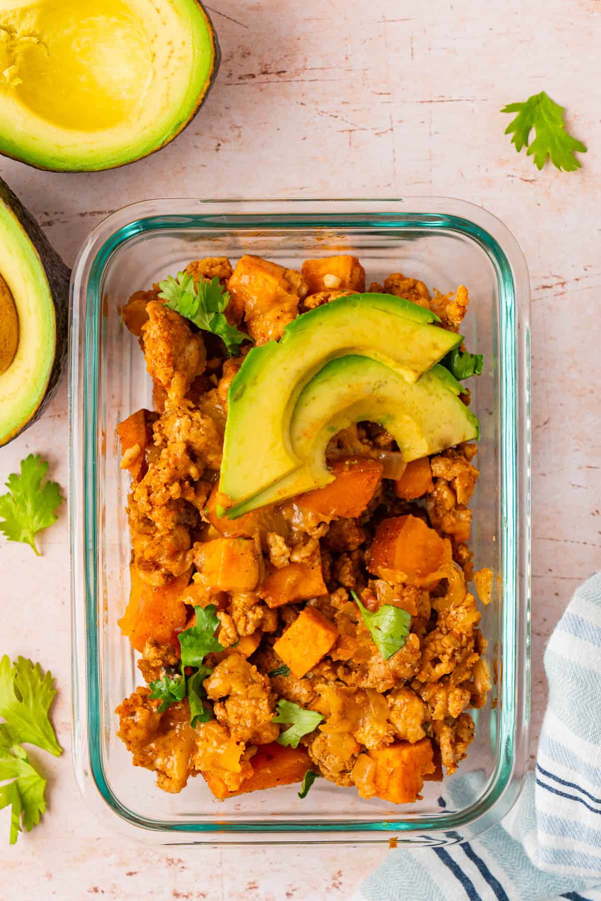 Sweet potato ground turkey skillet in a glass meal prep container topped with sliced avocado and fresh cilantro.