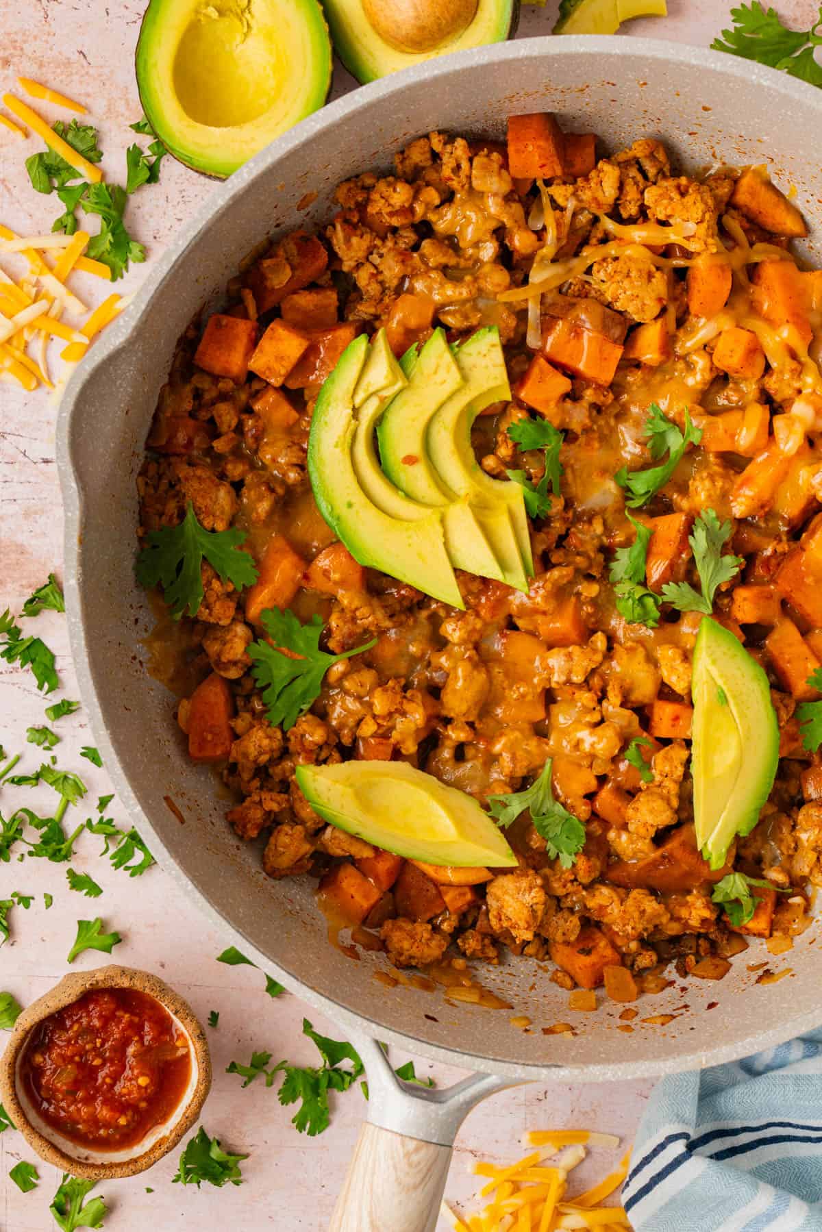 Ground turkey sweet potato skillet topped with melted cheese, sliced avocado, and fresh cilantro in a large white skillet.